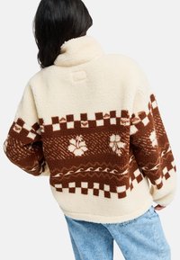 Pullover in pile color crema e marrone, con un motivo a quadri, dettagli floreali e un colletto alto con una finitura morbida e testurizzata.