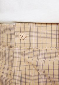 Vaaleankeltaiset housut, joissa on ruutukuosi tummansinistä ja valkoista. Varustettu napituksella ja hillityllä tekstuurilla.