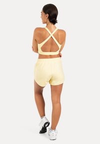Femme debout, dos tourné, portant un soutien-gorge de sport jaune pâle à dos croisé, un short assorti, des baskets blanches, sur un fond clair uni.