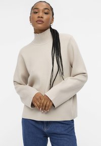 Beige strikkegenser med høy krage, løs passform og lange ermer; kombinert med blå jeans. Glatt tekstur, minimal detaljering.