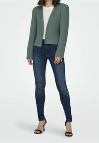 Blazer court texturé vert porté sur un haut blanc, associé à un jean slim bleu foncé et des sandales à talons noires.