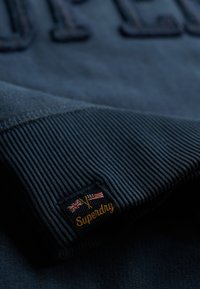 Manga acanalada oscura con un parche del logo de Superdry que muestra las banderas británica y americana sobre un fondo de tela oscura.