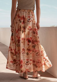 Fließender Maxirock aus leichtem Stoff mit floralem Muster in Rot und Grün auf beigem Hintergrund. Elastischer Bund. Klassische flache Sandalen.