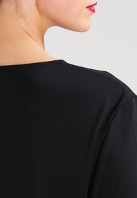 Käden ja olkapään kuva mustassa topissa, kirkkaan punaisella huulipunalla, vaaleanharmaata taustaa vasten.