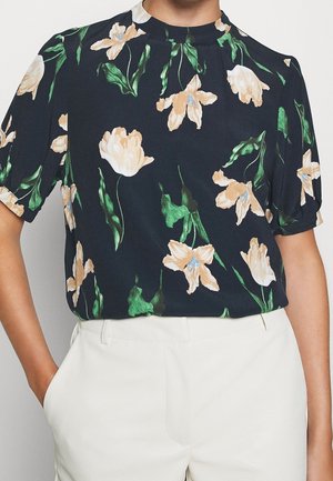 Vrouw draagt een donker bloemenblouse met beige bloemen en groene bladeren, gecombineerd met witte hoge taillebroeken, handen in de zakken.