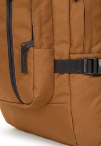 Hoek van een bruine stoffen rugzak met een met rits afgesloten zak en een zwarte riem met gesp. Geverfd materiaal met zichtbare naden en stiksels.