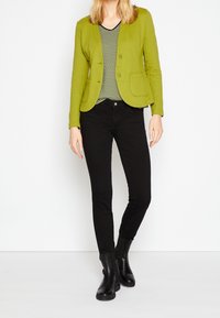 Blazer vert avec deux boutons et poches, superposé sur un débardeur à rayures noir et blanc à col en V. Jeans skinny noirs et bottines.