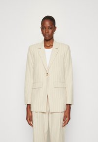 Beige pruhovaný blazer s jedním knoflíkem, klopami a předními kapsami, vyrobený z texturované látky, nošený přes bílou košili.