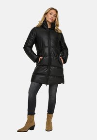 Zwarte gewatteerde lange jas met ritsluiting, hoge kraag en zijzakken. Draagt over donkere jeans en beige enkellaarzen. Soepele textuur.