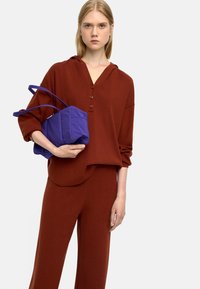 Brązowy dzianinowy hoodie z dekoltem w serek i trzema guzikami, zestawiony z pasującymi brązowymi spodniami, trzymający fioletową torbę z materiału z uchwytami.