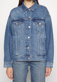 Chaqueta de denim en azul claro con un diseño clásico, que presenta dos bolsillos en el pecho, botones plateados y un corte ligeramente oversize.