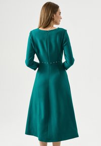 Teal midi šaty s dlhými rukávmi, okrúhlym výstrihom a priliehavým pásom. Funkcie ozdobné hroty pozdĺž pása a centrálne zadný zips.