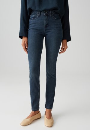 Vrouw draagt donkerblauwe skinny jeans, marineblauw shirt met lange mouwen en beige platte schoenen, staand tegen een effen lichte achtergrond.