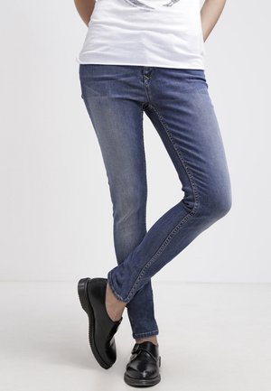 Personne portant un jean skinny bleu et des chaussures en cuir noir sans lacets, debout jambes croisées devant un fond blanc uni.