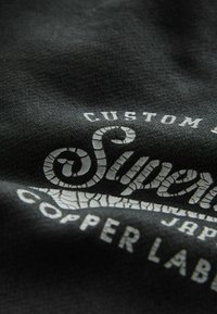 Schwarzes Stoff-T-Shirt mit weißem, rissigem Textlogo "Superdry" und "Copper Label" in kontrastierendem Druck, das subtile Textur und Details aufweist.