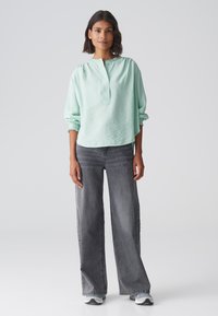 Blusa a maniche lunghe di un verde menta chiaro con una finitura testurizzata, dotata di un colletto con bottoni. Abbinata a jeans in denim grigio a gamba larga.