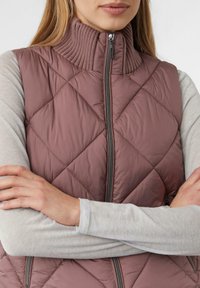 Femme les bras croisés portant un gilet matelassé rose poudré avec un col haut côtelé et une chemise à manches longues gris clair en dessous.