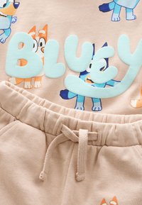 Sweat-shirt beige clair avec des illustrations ludiques de chiens et le mot "BLUEY" en lettres aqua douces ; assorti d'un pantalon de survêtement beige avec taille à cordon.