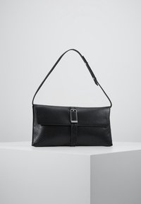 Černá texturovaná kožená taška přes rameno s klopou a detaily s přezkou, umístěná na bílé podstavě na pozadí jednoduchého šedého pozadí.