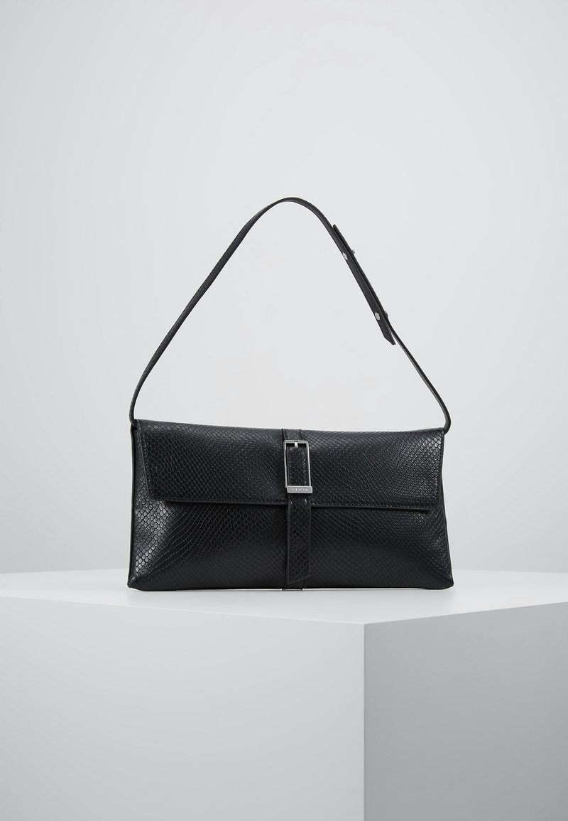 Černá texturovaná kožená taška přes rameno s klopou a detaily s přezkou, umístěná na bílé podstavě na pozadí jednoduchého šedého pozadí.