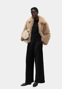Sztuczny futrzany płaszcz w jasnym beżu, strukturalny czarny kombinezon, czarne płaskie buty oraz torebka w kolorze kremowym z węzłem. Poza stojąca na jednolitym tle.