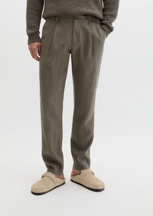 Osoba ubrana w oliwkowe spodnie, beżowe buty wsuwane i brązowy dziergany sweter, stoi z jedną ręką w kieszeni.