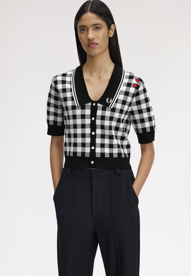 Femme portant une chemise à carreaux noire et blanche avec un col noir, des pièces en forme de cœur rouges sur l'épaule, et un pantalon noir les mains dans les poches.
