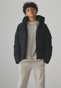 Sort puffervest med hætte, der har et quiltet design. Båret over en lys grå sweatshirt og beige sweatpants med subtil tekstdetalje.
