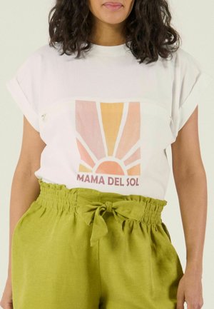 Vrouw draagt een wit T-shirt met opgerolde mouwen en een kleurrijke zonnenprint met de tekst "MAMA DEL SOL", gecombineerd met een groene high-waisted paperbagbroek.