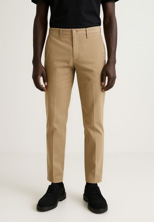 Beige Stoffhosen mit schmaler Passform, flachem Bund und zwei Seitentaschen. Kombiniert mit schwarzen Schuhen und einem schwarzen Hemd für einen Kontrast.