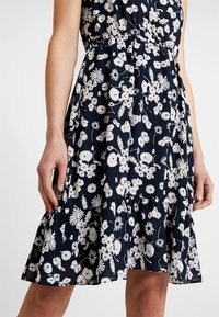 Femme portant une robe bleu marine avec un motif floral blanc et un ourlet volanté, montrée de la mi-cuisse à la taille sur un fond uni.