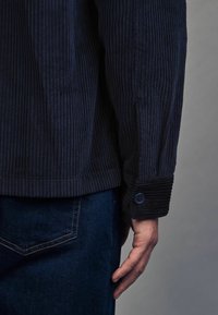 Laivastonsininen corduroy-takki, jossa on ribbetrakatoitu pinta; siinä on napilla kiinnitetyt hihansuut ja suora leikkaus. Päällä tummat farkut.