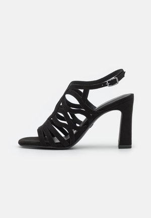 Czarne zamszowe sandały na wysokim obcasie mają strappy design z okrągłymi wycięciami, pasek na kostkę z klamrą oraz kwadratowy obcas.