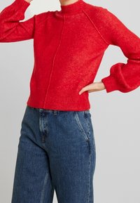Roter, strukturierter Pullover mit hohem Kragen, Raglanärmeln und gerippten Bündchen. Kombiniert mit hochgeschnittenen blauen Jeans.