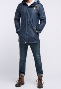 Navy waterproof jacket with a hood, zip front, and buttons. Features a grey interior lining and side pockets, paired with dark jeans and brown boots.