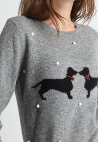 Pull gris avec des silhouettes de chiens noirs se faisant face, portant des colliers rouges, décoré de petits pompons blancs autour du motif.
