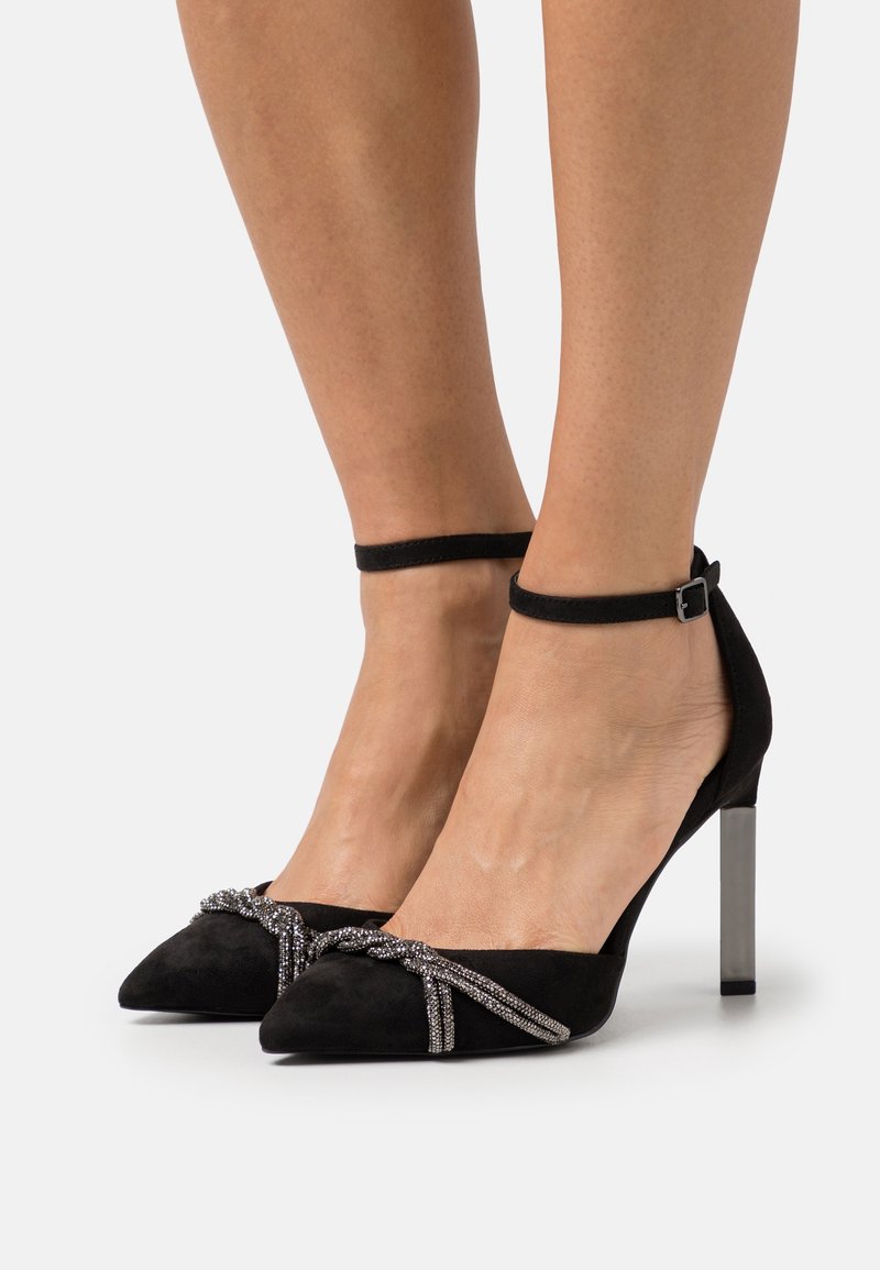 Chaussures à talons hauts en daim noir avec un bout pointu, un design unique de nœud orné d'accents métalliques, et un talon métallique élancé. Bride de cheville incluse.