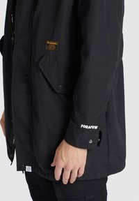 La main d'un homme repose le long du corps, portant une veste noire avec une poche à bouton, une étiquette "Khuogenuine" sur la poitrine et "FORAFEW" brodé sur le poignet.