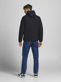 Giacca a vento nera con cuciture orizzontali e cappuccio blu navy, abbinata a jeans blu e sneakers con dettagli verde, vista da dietro.