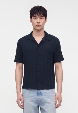 Jeune homme aux cheveux courts noirs portant une chemise sombre à manches courtes boutonnée et un jean bleu clair, debout devant un fond blanc uni.