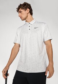 Nike polo shirt i lys grå, med et blomsterdesign, knaplukning og et sort logo på brystet. Korte ærmer og afslappet pasform.