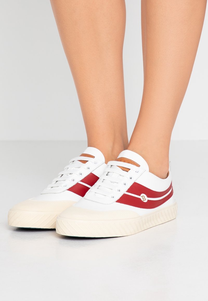 Baskets blanches avec des bandes rouges et des semelles beige portées pieds nus sur un fond clair uni.