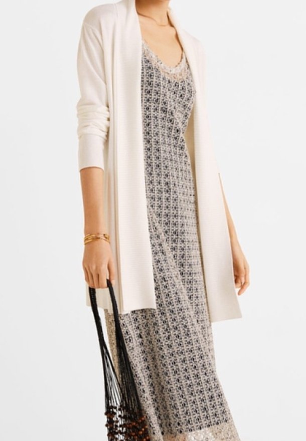 Femme portant une robe maxi sans manches à motifs avec un cardigan blanc ouvert, tenant un sac à cordon noir avec des perles en bois.