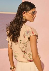 Blusa floreale in morbido tessuto crema con maniche arricciate e bordo in pizzo, caratterizzata da vivaci motivi floreali e abbinata a pantaloni chiari.