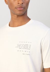 Λευκό βαμβακερό t-shirt με μαύρο κείμενο, περιλαμβάνοντας συντεταγμένες και ένα ενθαρρυντικό απόφθεγμα. Κλασική στρογγυλή λαιμόκοψη και κοντά μανίκια.