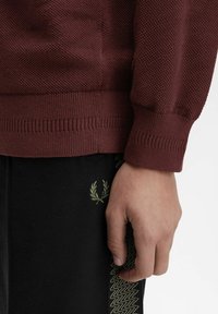 Primo piano di una mano accanto a un polsino in maglia bordeaux e a pantaloni neri con un piccolo logo a ricamo di una corona di alloro e una striscia a motivo.