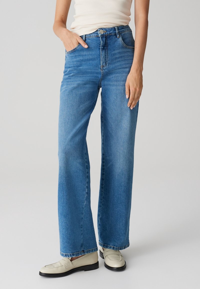 Femme portant un jean bleu à jambes larges, des mocassins blancs et un top blanc sans manches, main dans la poche, debout devant un fond uni.