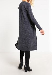 Lång grå stickad klänning med långa ärmar och fladdrande fåll. Bärs över svarta leggings och tillsammans med svarta klackade ankelskor.