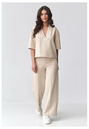 Kobieta z długimi falowanymi włosami, ubrana w luźną beżową bluzkę z dekoltem w serek i pasujące spodnie o szerokich nogawkach, idzie do przodu w białych trampkach na białym tle.