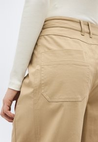 Μπεζ παντελόνι με μεγάλη πίσω τσέπη, συνδυασμένο με κρεμ ριμπ μακρυμάνικη μπλούζα, που δείχνει κοντινό πλάνο της μέσης και του χεριού.